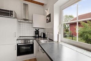 Et køkken med hvide skabe, en stålkogeplade, en dobbeltvask og et vindue med udsigt til et rødt hus.