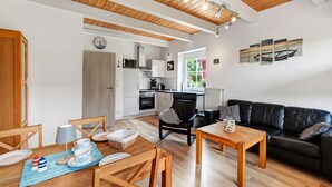 A modern kitchen with white cabinets, a dining area with a wooden table and chairs, a black leather sofa, and a view of the outdoors. - Holiday Home Little Lighthouse-Backbord Terrace, Garden, Wi-Fi, Bike Garage, EV Charging (Fehmarn)