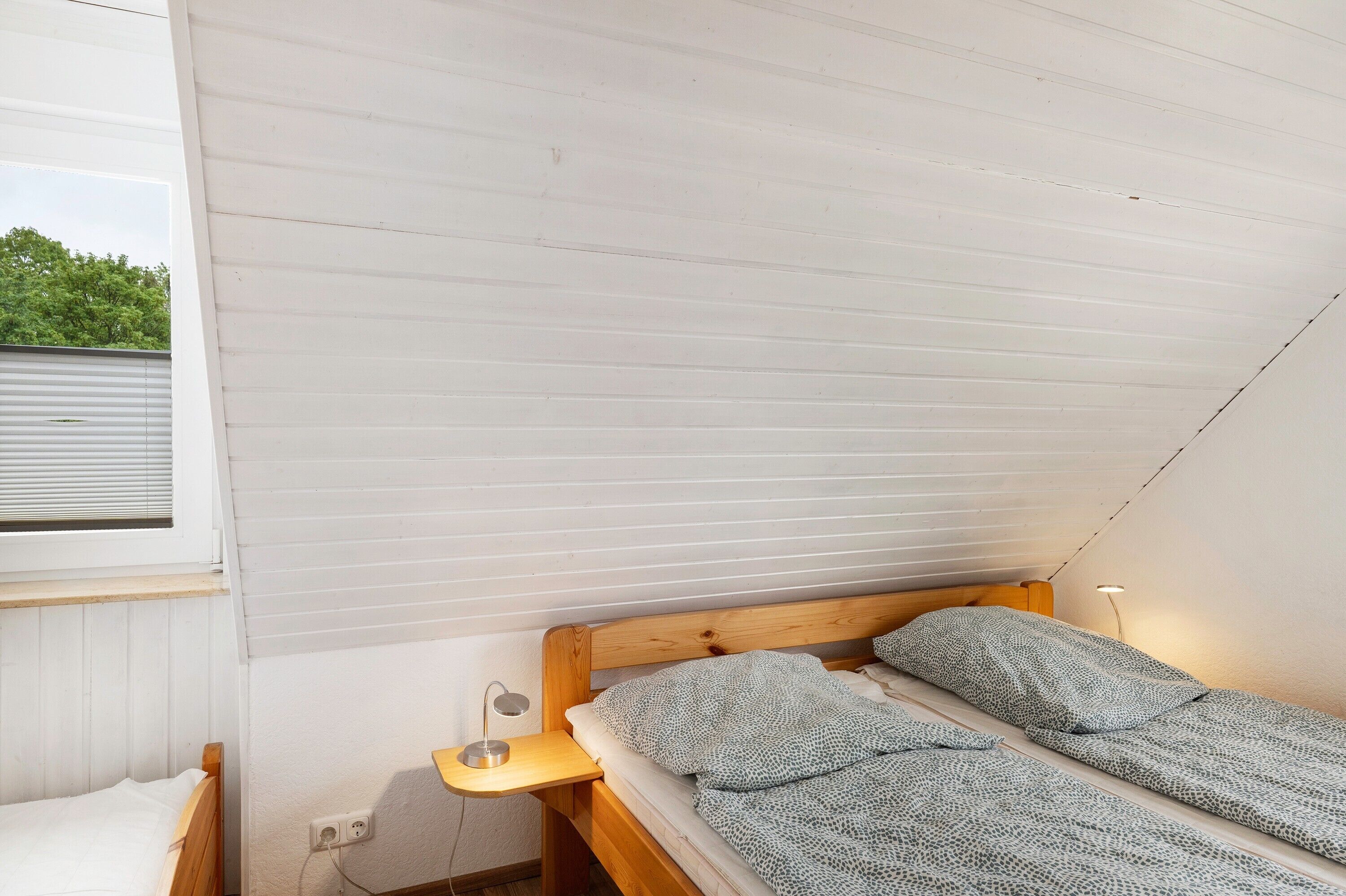 Ein Einzelbett mit Holz-Kopfteil, ein Nachttisch mit Lampe und ein Fenster mit Jalousien.