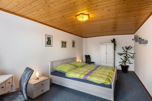 Une chambre avec un plafond en bois, un lit recouvert d’une couverture verte et violette, une table de chevet avec une lampe, une chaise, une armoire et une plante en pot.