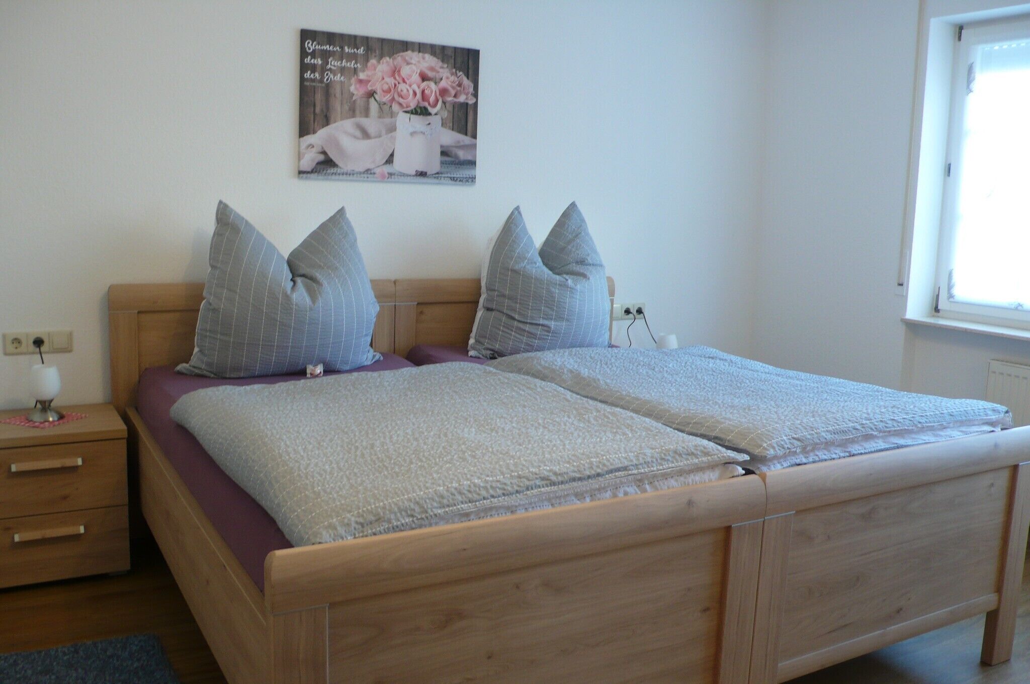 Ein Doppelbett mit Holzrahmen, zwei Nachttische, ein Nachtschränkchen, eine Wandsteckdose, ein Fenster mit Jalousien und ein Poster an der Wand.