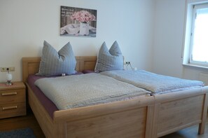 A double bed with a wooden frame, two bedside tables, a nightstand, a wall-mounted power outlet, a window with blinds, and a poster on the wall.
