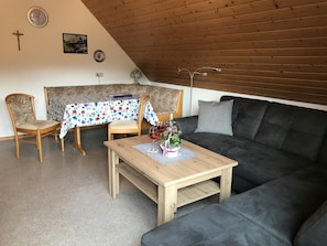 A cozy living room with a wooden ceiling, a sofa, two chairs, a table with a floral tablecloth, and a clock on the wall.
