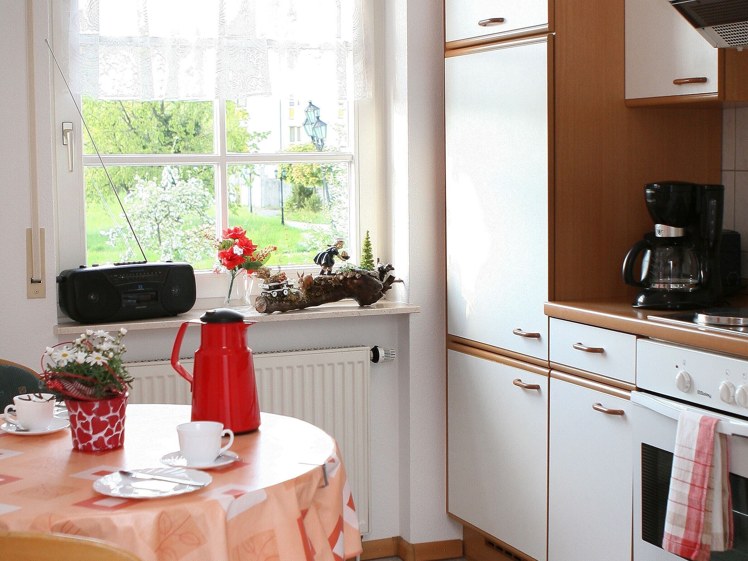 Una cocina con ventana, un radiador, una cafetera y una mesa con un hervidor rojo.