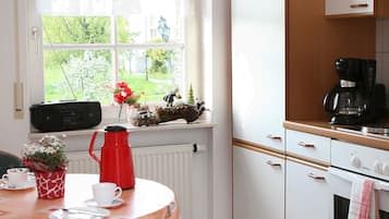 Una cocina con ventana, un radiador, una cafetera y una mesa con un hervidor rojo.