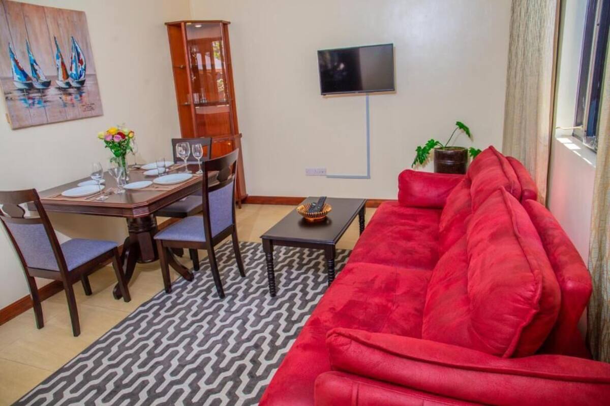 A dining area with a wooden table set for four, a red sofa, a patterned rug, a television on the wall, and a cabinet.