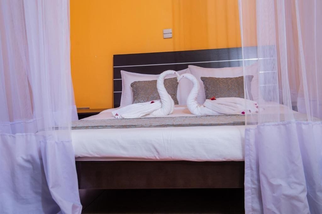 Una cama con sábanas blancas, dos toallas con forma de cisne, un cabecero negro y una pared naranja lisa.