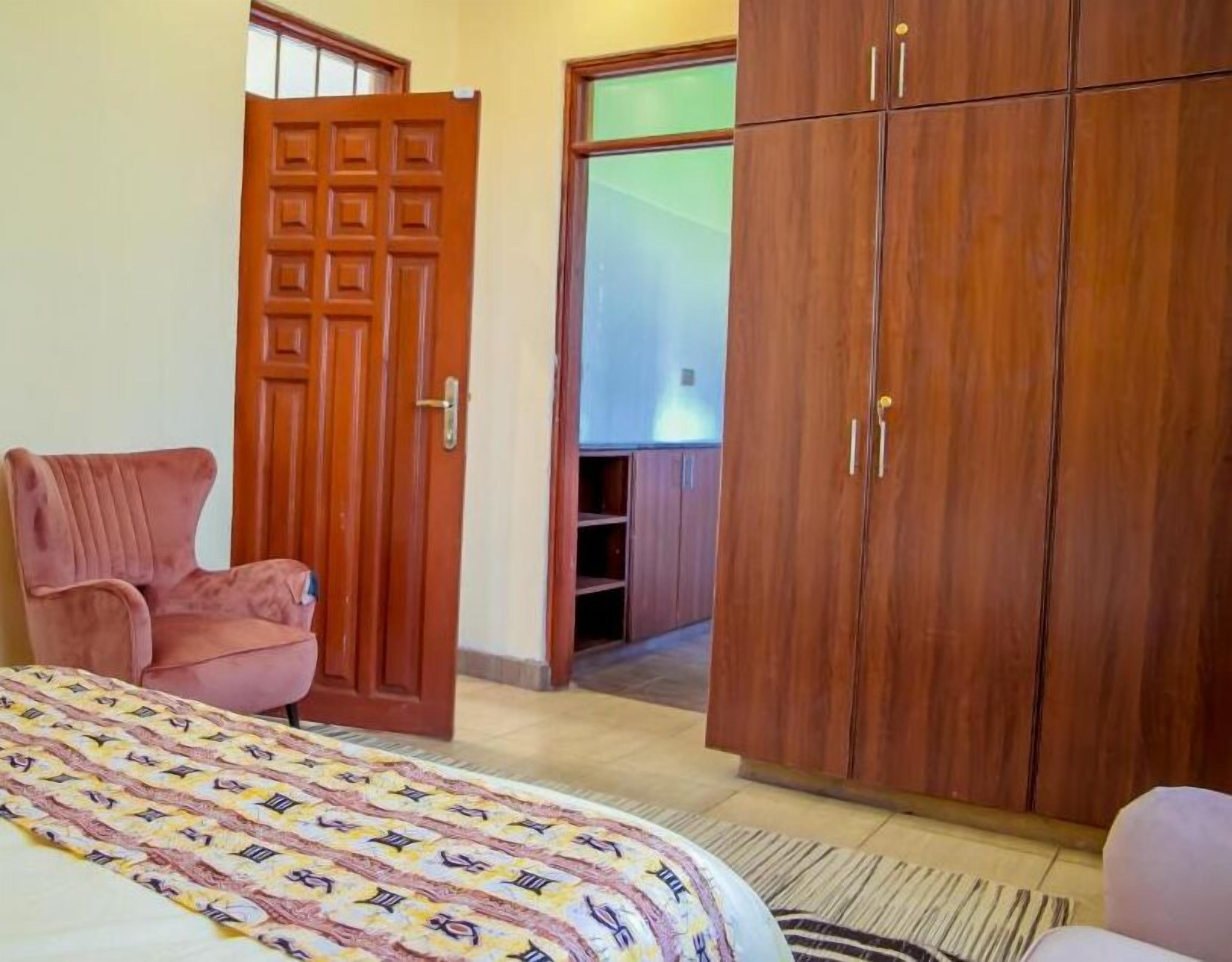 A bedroom with a bed, a pink chair, wooden wardrobe, and a door leading to a balcony.