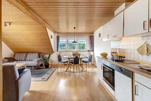 A modern kitchen with wooden beams, a dining area with a round table and chairs, and a view of greenery outside.