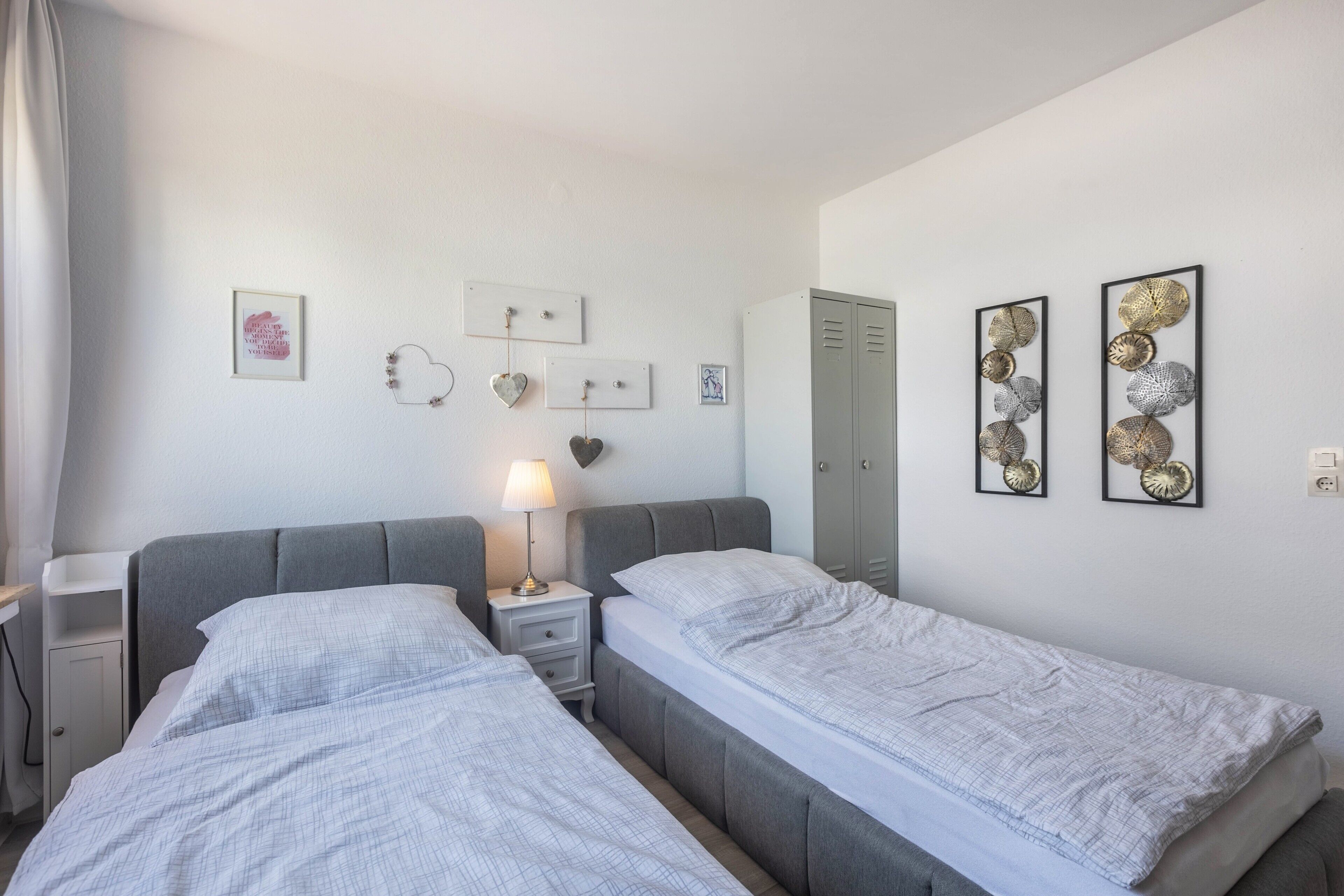 Una camera d'albergo con due letti, un comodino con una lampada, un armadio e decorazioni a parete tra cui quadri incorniciati e un ornamento a forma di cuore.