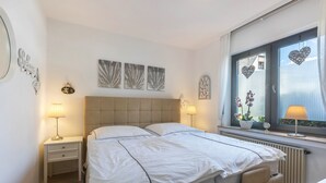 A bedroom with a bed, two bedside tables, a window with heart-shaped decorations, and wall art.