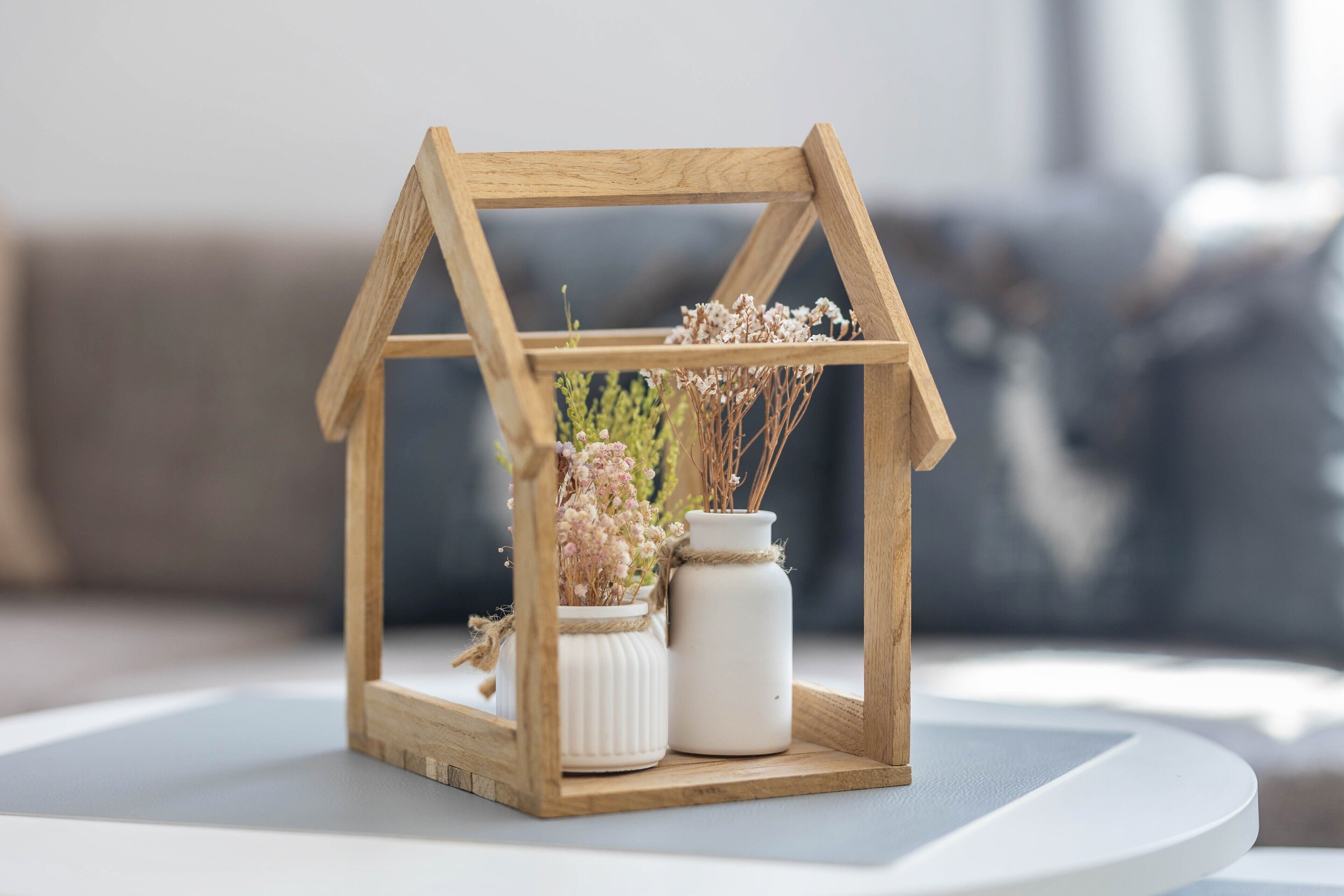 Um porta-retrato em forma de casa de madeira com flores secas e um vaso branco sobre uma mesa branca.