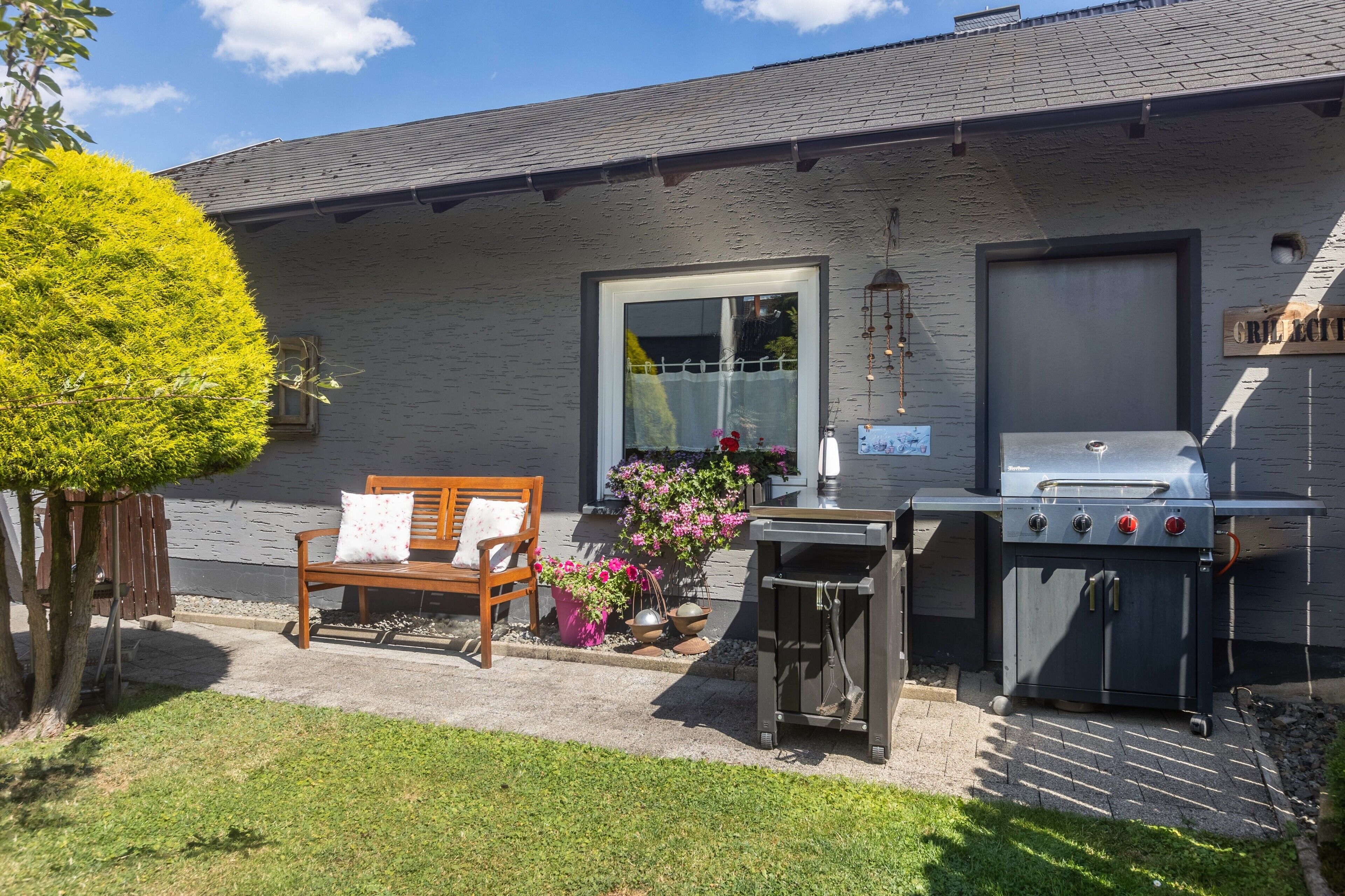 Eine Terrasse mit einer Holzbank, einem Grill und Kübelpflanzen.