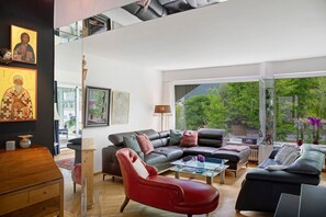 A spacious living room with a large sofa, a red armchair, and a glass coffee table. There are framed pictures on the wall and a view of greenery outside. - Haus Gutschi with Mountain Views, Large Garden and Wi-Fi (Gemeinde Imst)