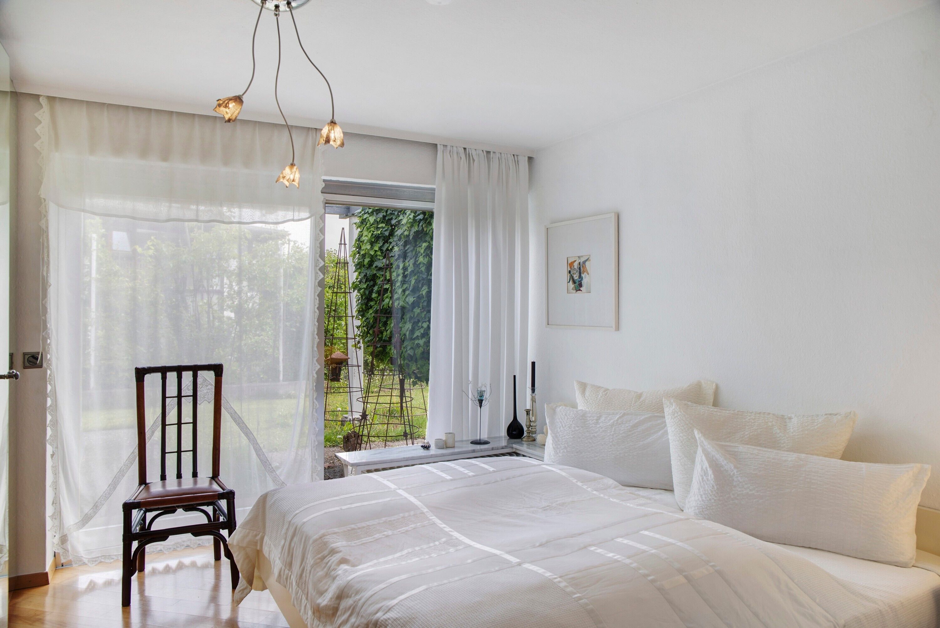 Un dormitorio con cama, una silla, un ventanal con cortinas traslúcidas y vistas a zonas verdes.
