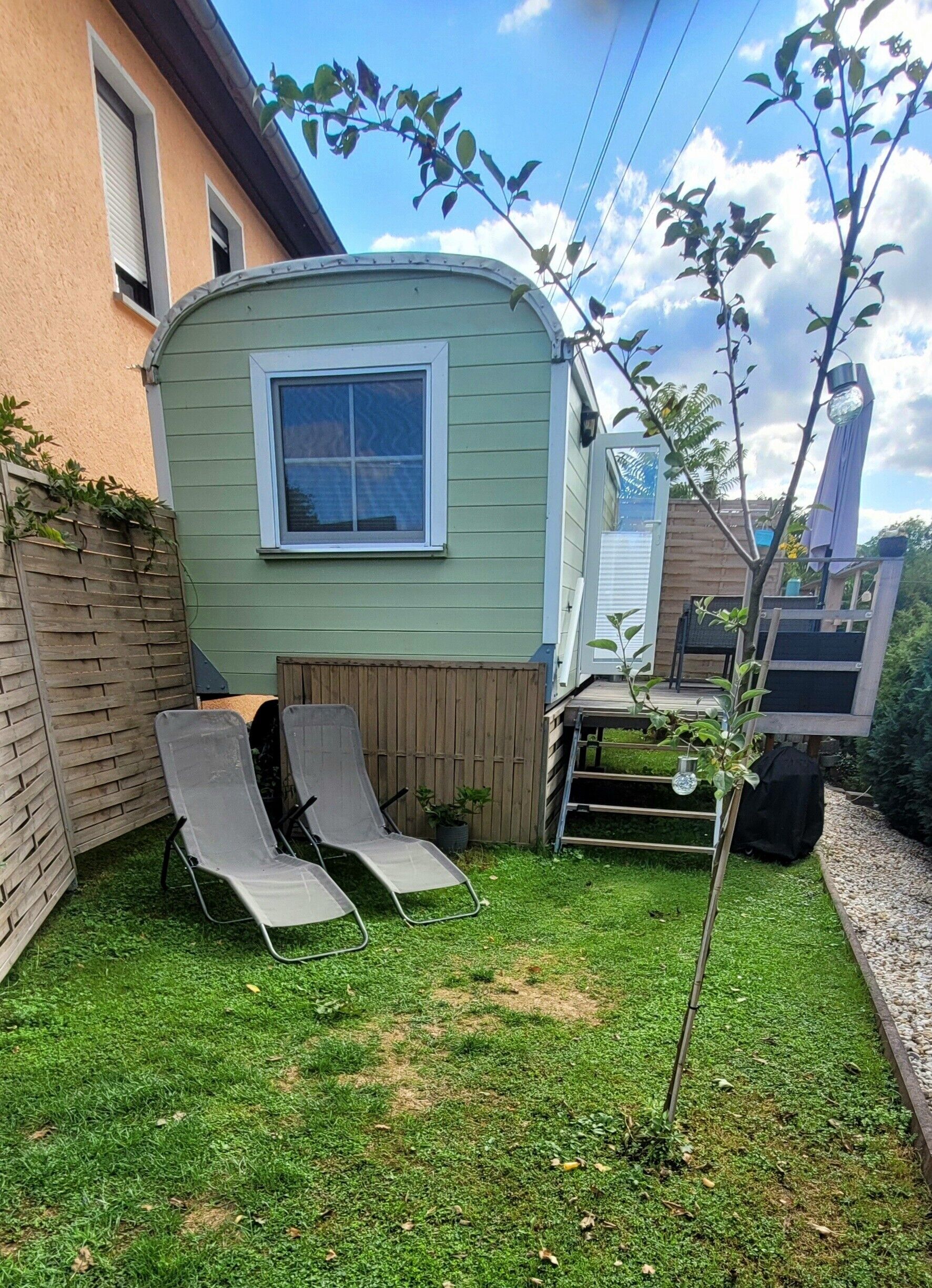 Une petite construction à un seul étage, dotée d’une fenêtre, d’une clôture en bois et de deux chaises longues dans une cour verdoyante.