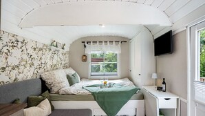 A compact bedroom with a bed, a small desk, a television, and a window with a view of greenery. - Tiny Home 'Amselnest' with Lake View, Private Terrace, and Wi-Fi (Oelsnitz/Vogtl.)