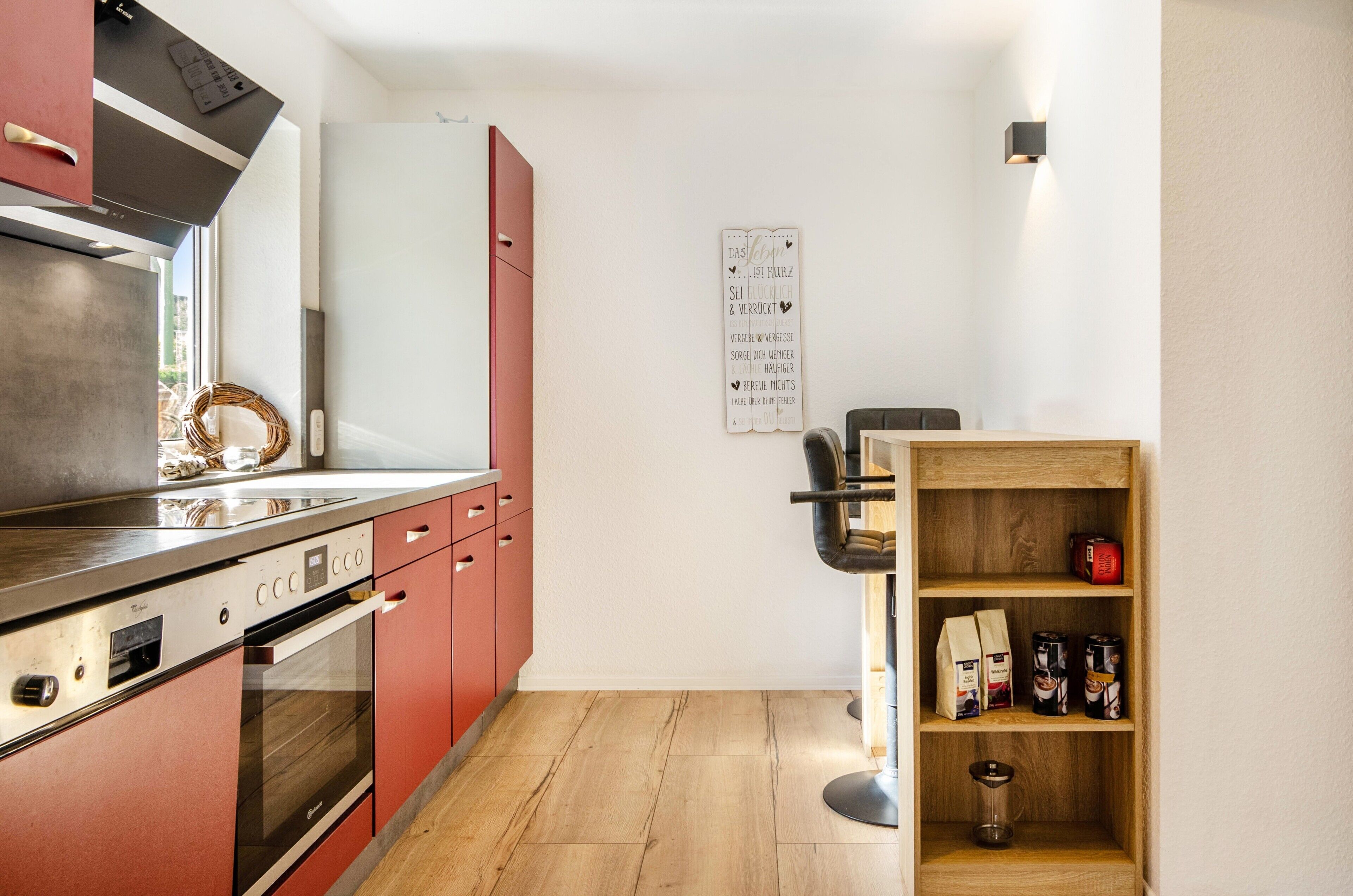 A compact kitchen with modern appliances, a wooden shelf with kitchen items, and a wall-mounted sign.