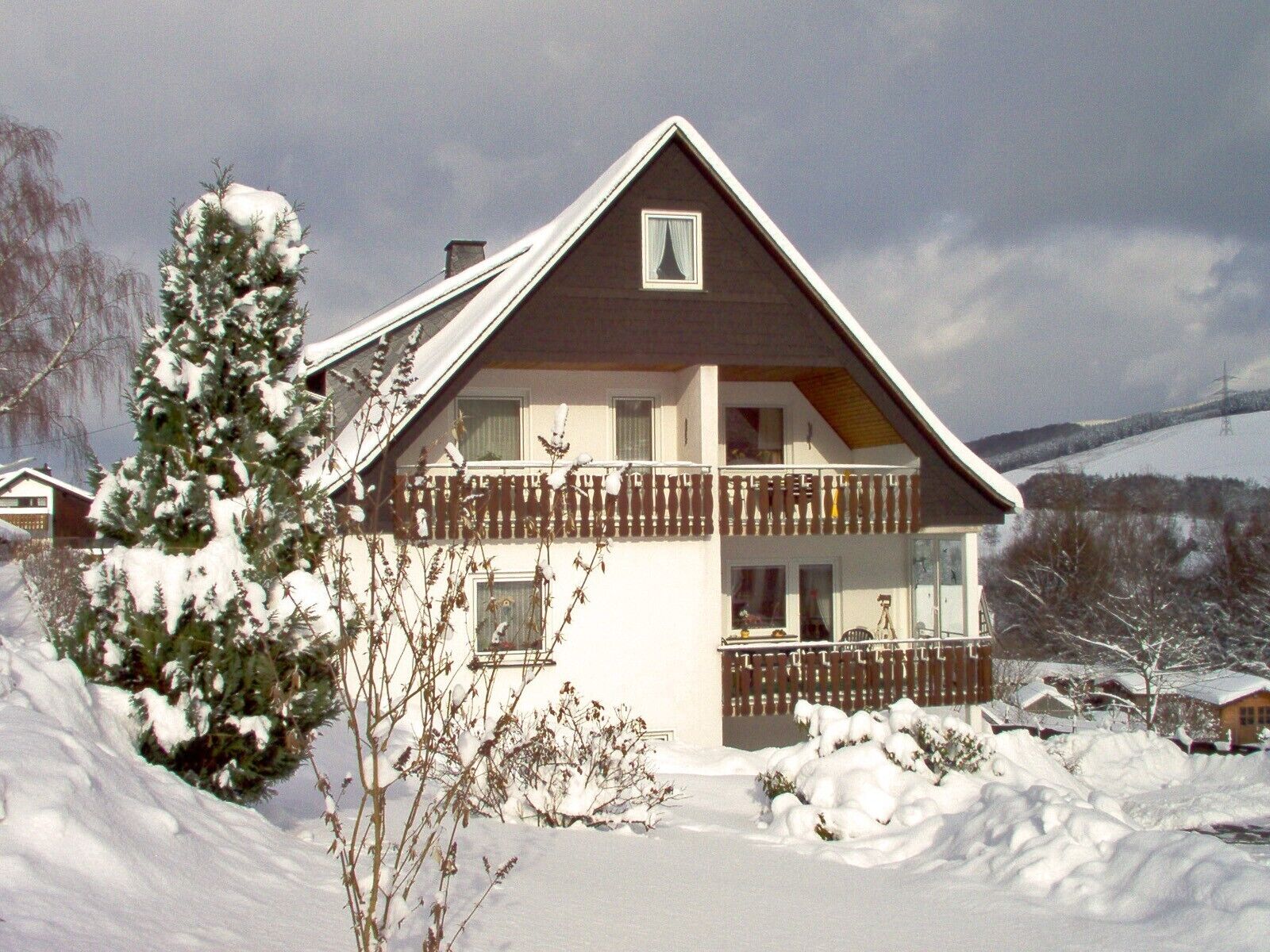 A two-story house with a balcony, surrounded by snow-covered ground and trees.