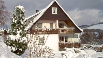 Una casa de dos pisos con balcón, rodeada de terreno y árboles cubiertos de nieve.