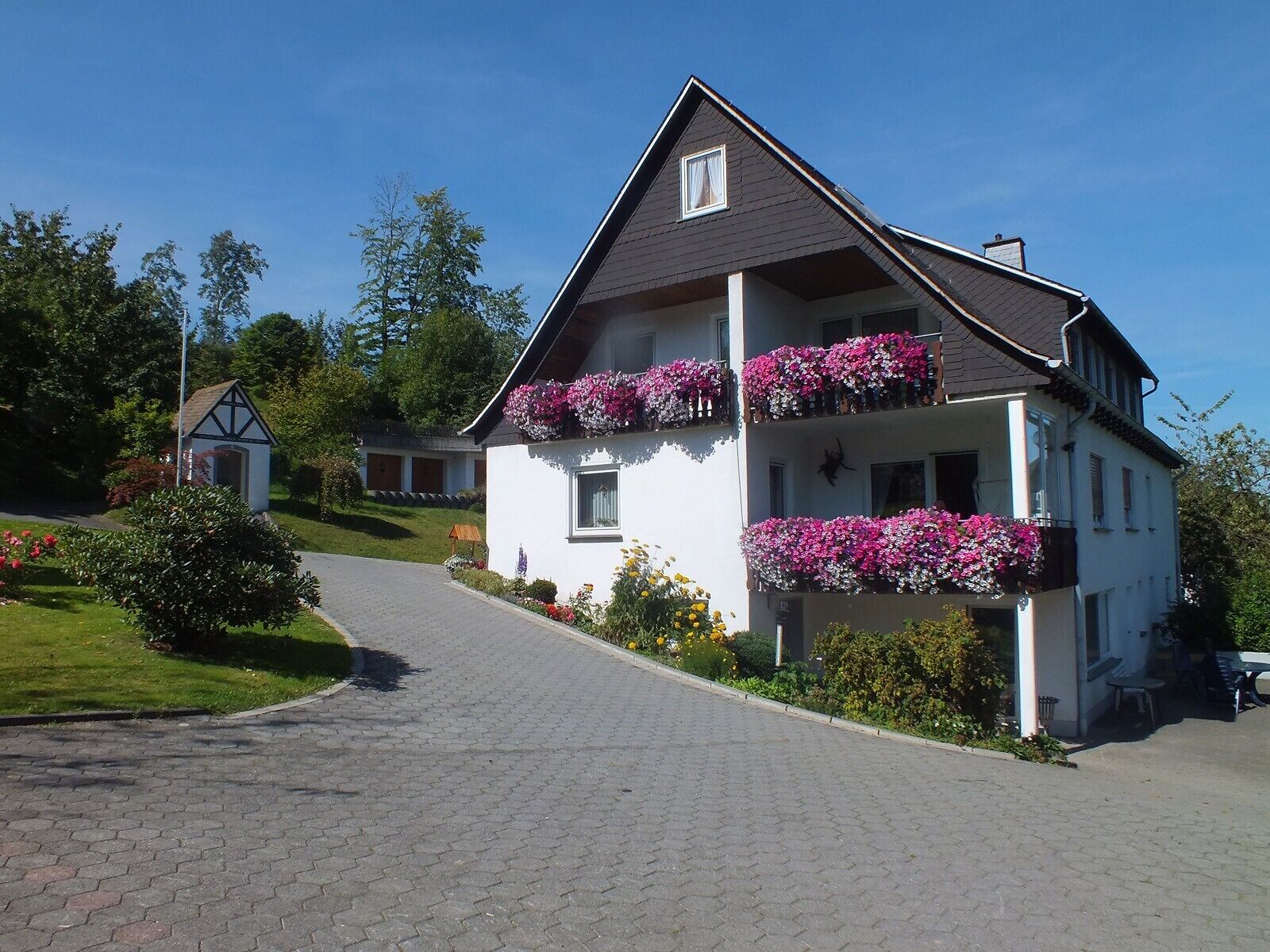 Una casa de dos pisos con balcones decorados con flores, un camino de entrada pavimentado y una pequeña construcción al fondo.