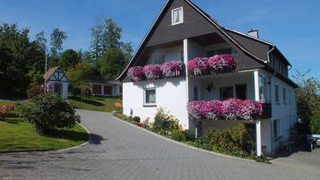 Una casa de dos pisos con balcones decorados con flores, un camino de entrada pavimentado y una pequeña construcción al fondo.