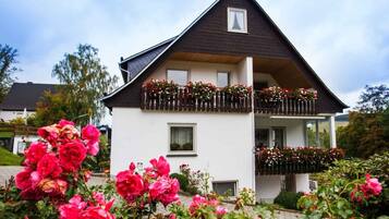 Ein zweigeschossiges Haus mit dunklem Dach und weißen Wänden, mit einem Balkon mit Blumenkästen und einem kleinen Fenster.