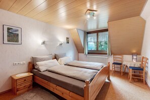 Ein Schlafzimmer mit einem hölzernen Bett, Nachttischen, einem kleinen Tisch mit Stühlen, einem Fenster mit Vorhängen und einer Heizung.