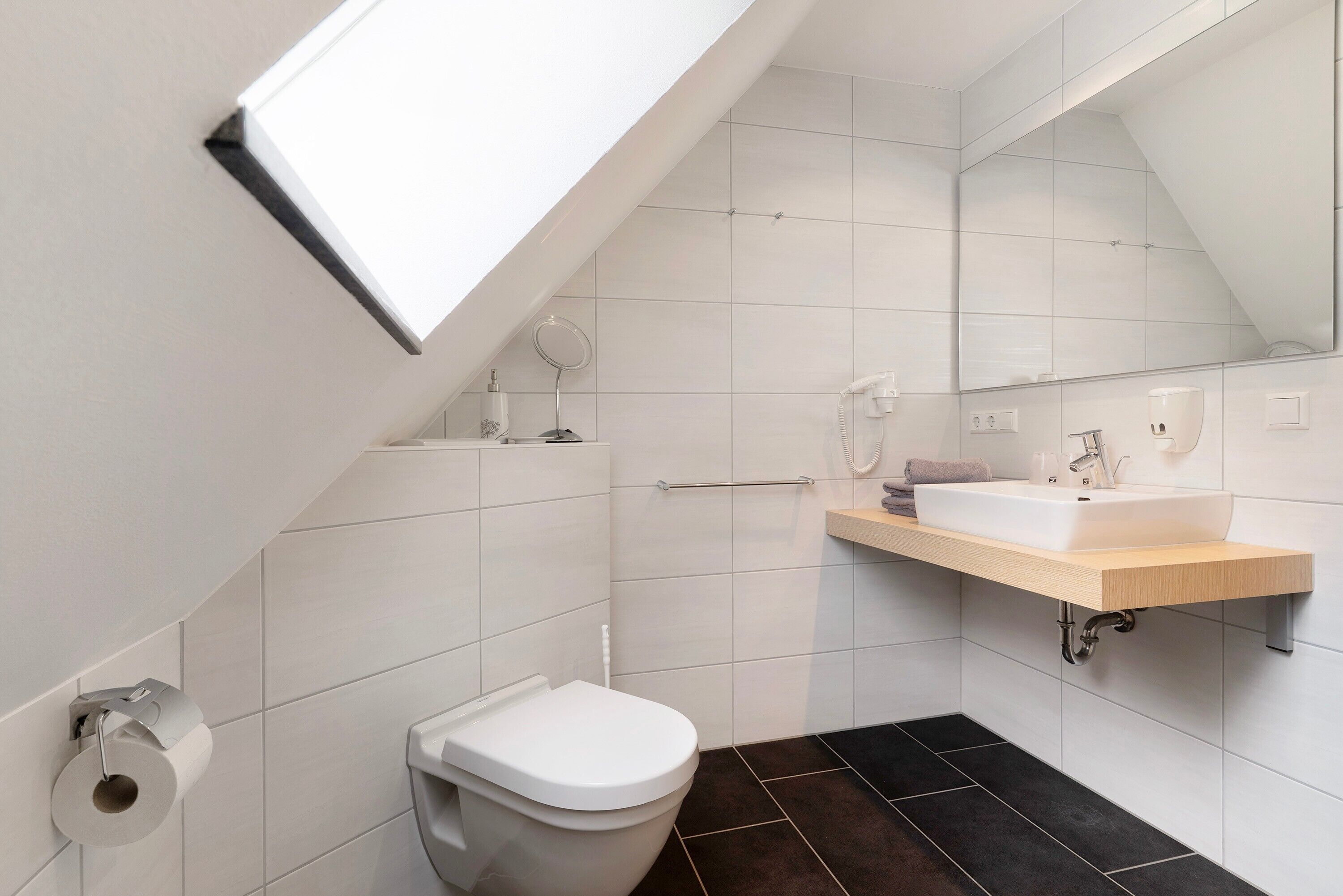 A modern bathroom with a skylight, white tiles, a toilet, a sink, and a mirror.