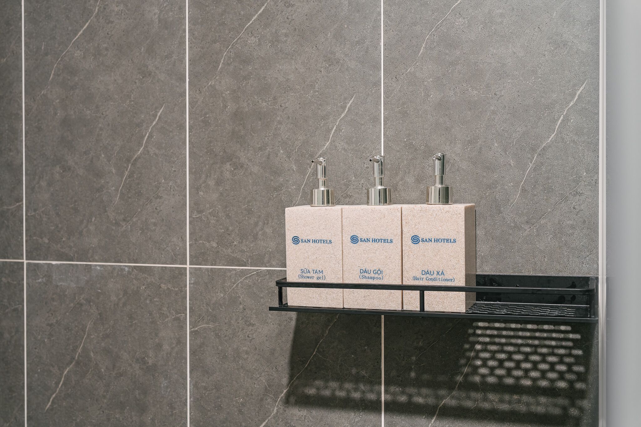 A bathroom with a marble wall, a shelf holding three bottles labeled 'SAN HOTELS' with 'SUA TAM' (shower gel), 'DAU GÒI' (shampoo), and 'DAU XÁ' (hair conditioner).