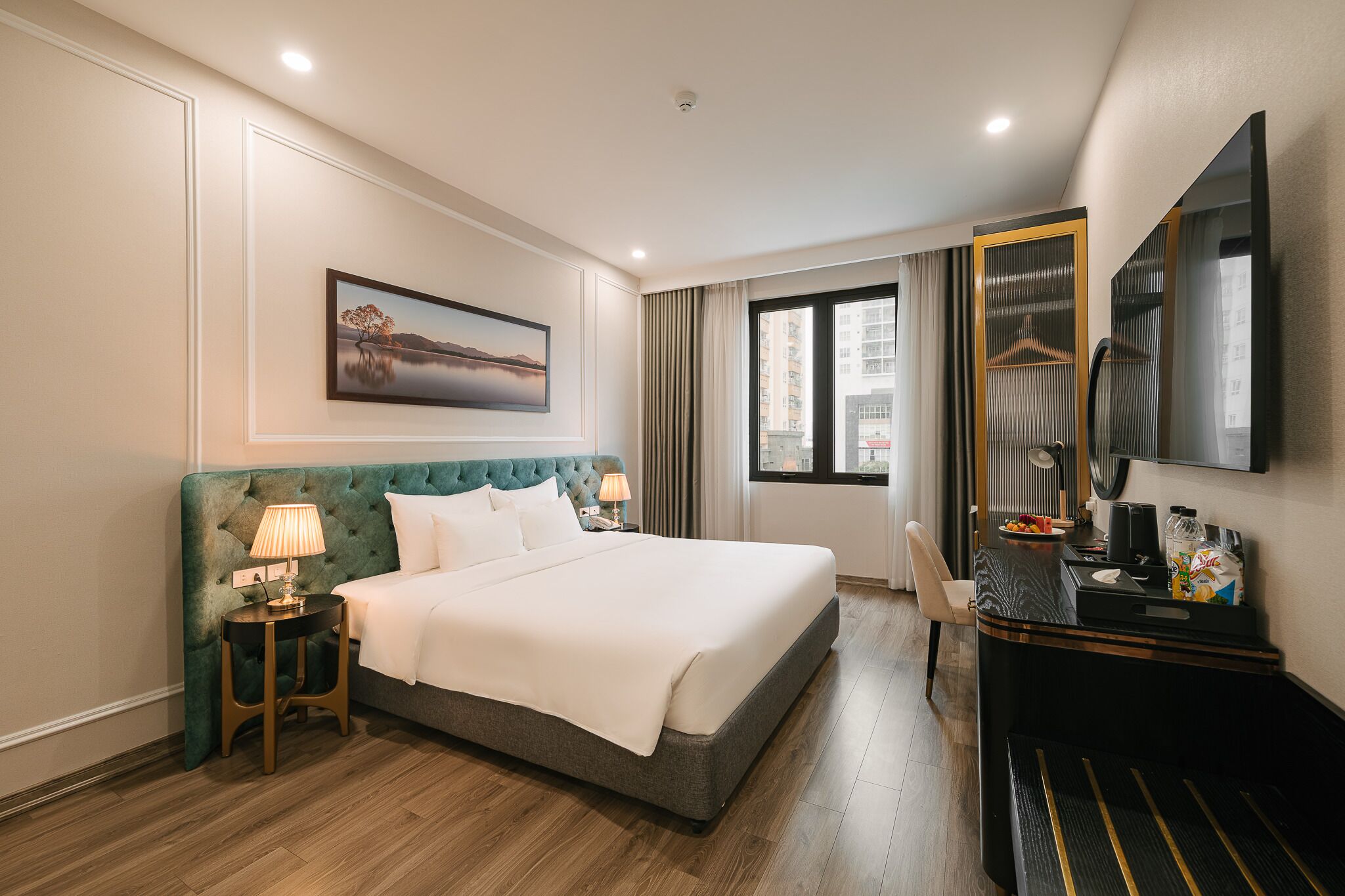 A hotel room with a large bed, a desk with a chair, a window with curtains, a painting on the wall, and a mirror on the wall.