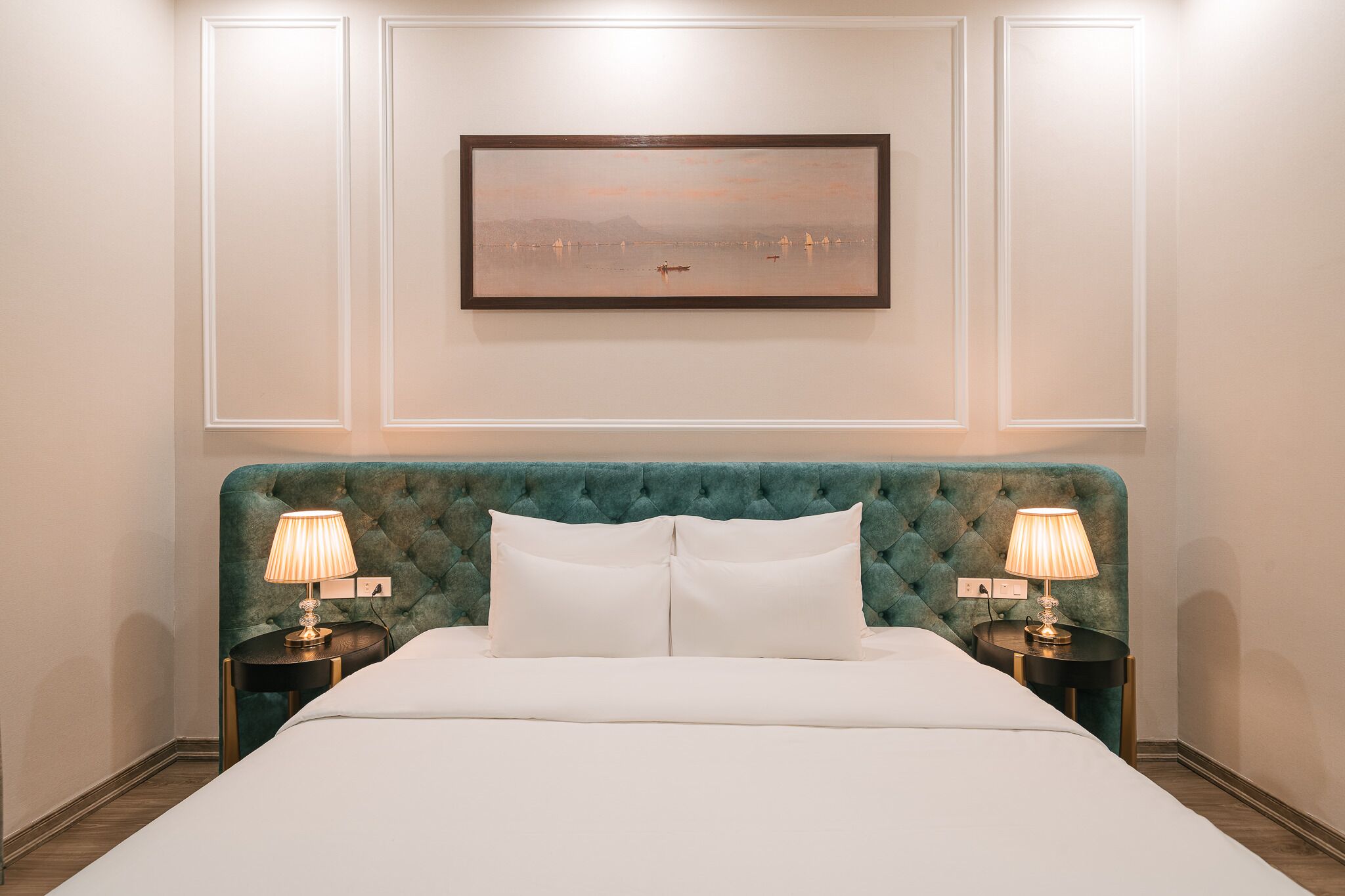 Une chambre d’hôtel avec un grand lit, deux tables de chevet avec des lampes, un tableau représentant un paysage encadré au mur et une tête de lit recouverte d’un tissu vert.