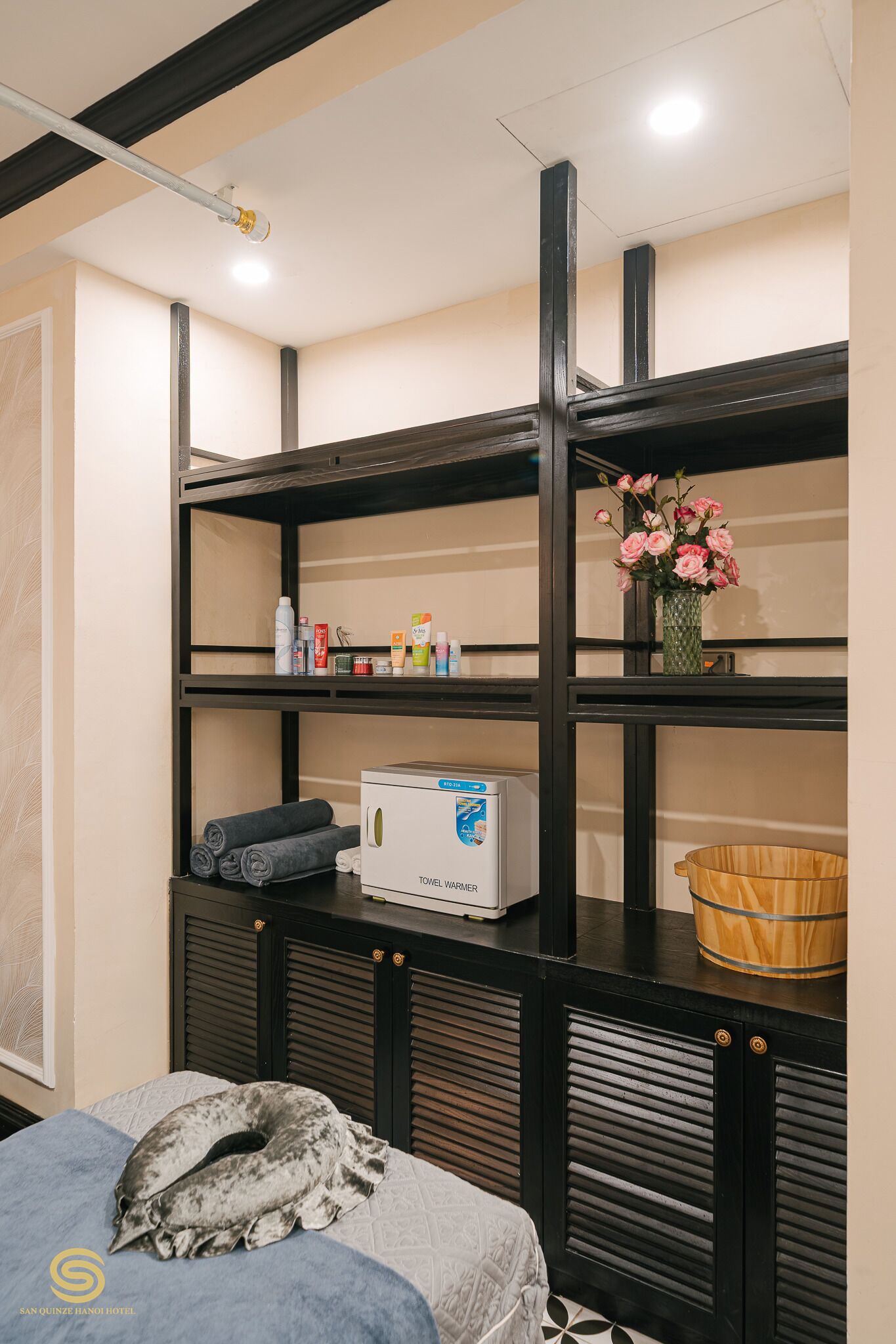 A hotel room with a bed, a towel warmer, a shelf with toiletries, and a vase with flowers.