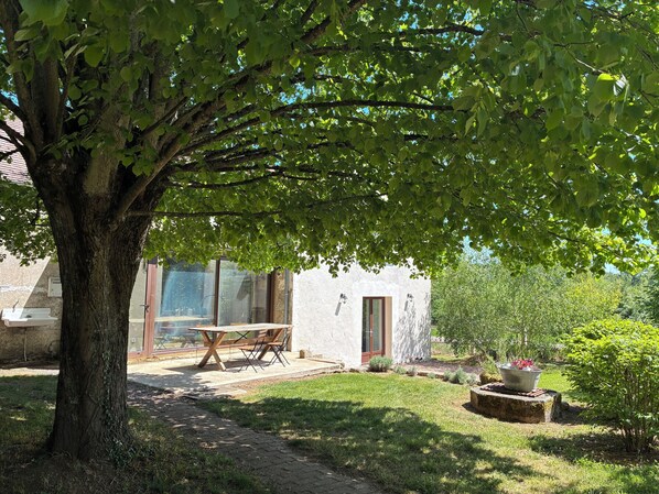 A tree-lined outdoor area with a table and chairs, a white building with large windows, and a well-maintained lawn.