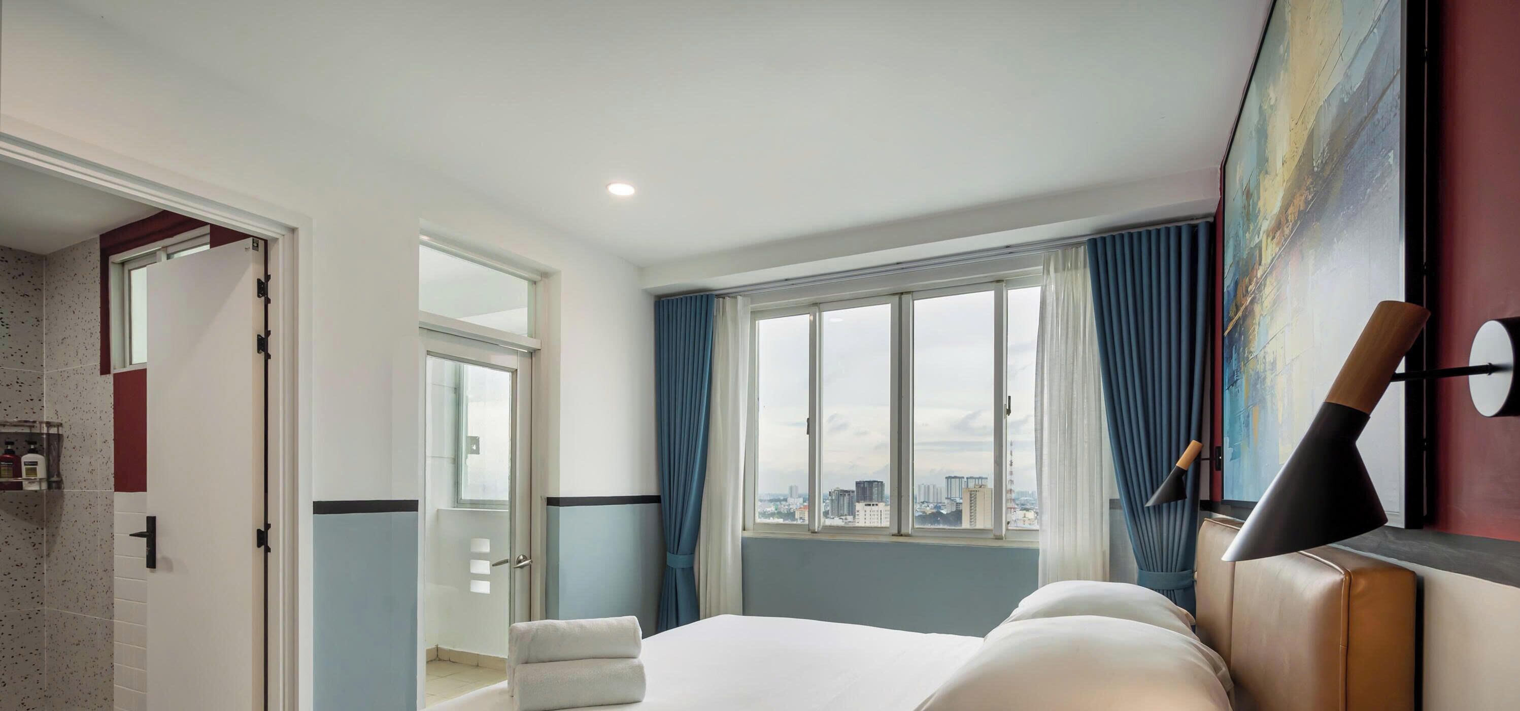 A hotel room with a bed, a window with a city view, a door leading to a bathroom, and a painting on the wall.