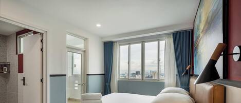 A hotel room with a bed, a window with a city view, a door leading to a bathroom, and a painting on the wall.
