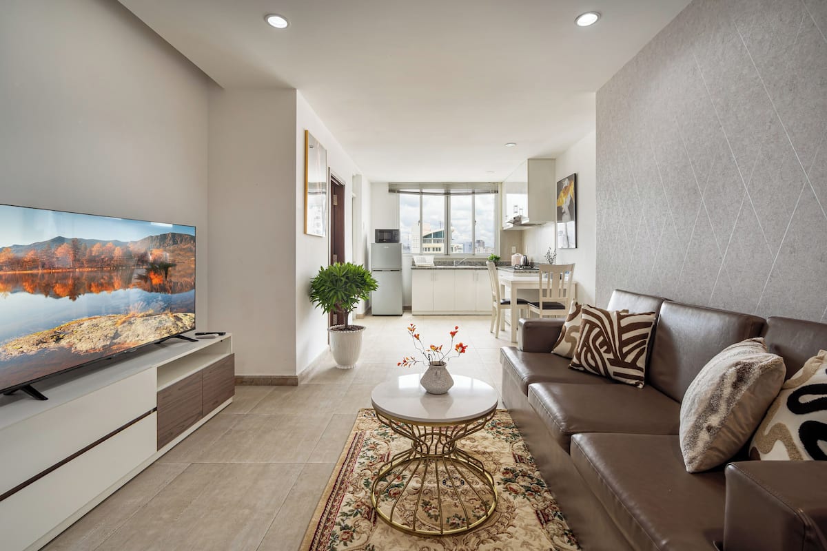 A modern living room with a flat-screen TV, a brown leather sofa, a round coffee table, and a large window.