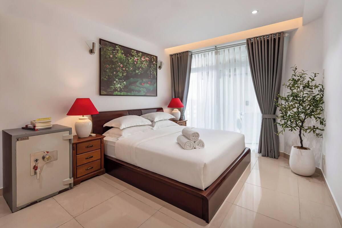 A hotel room with a bed, bedside lamps, a safe, a nightstand, a potted plant, and a painting on the wall.