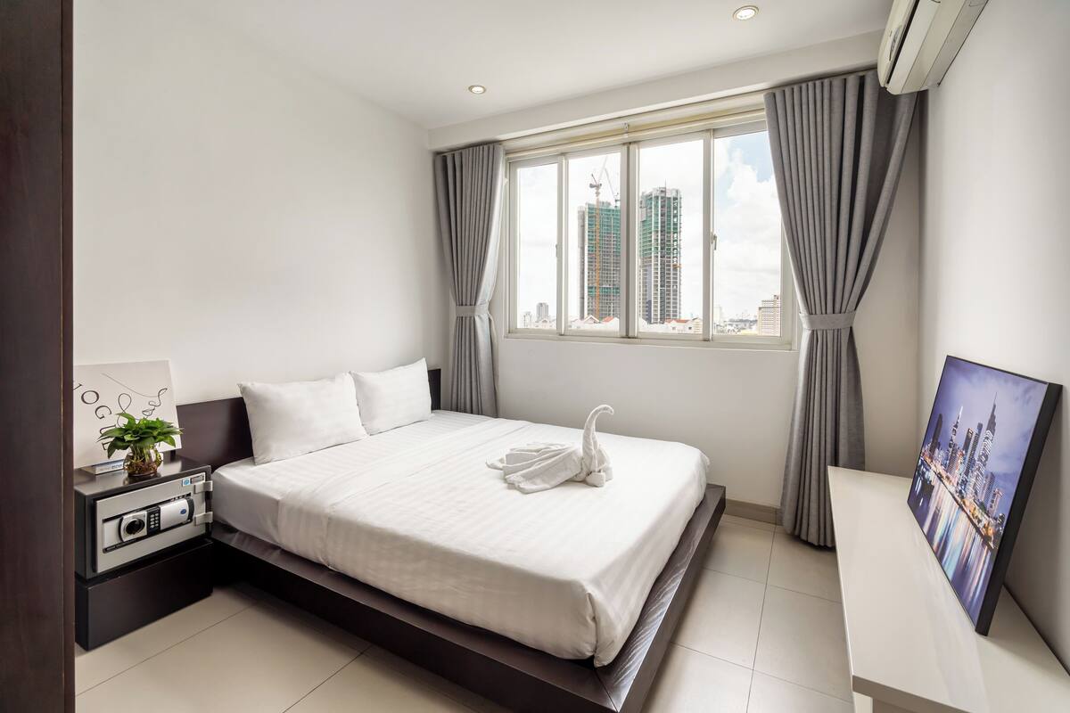 A modern hotel room with a large bed, a bedside table with a plant, a wall-mounted air conditioner, and a cityscape view through the window.