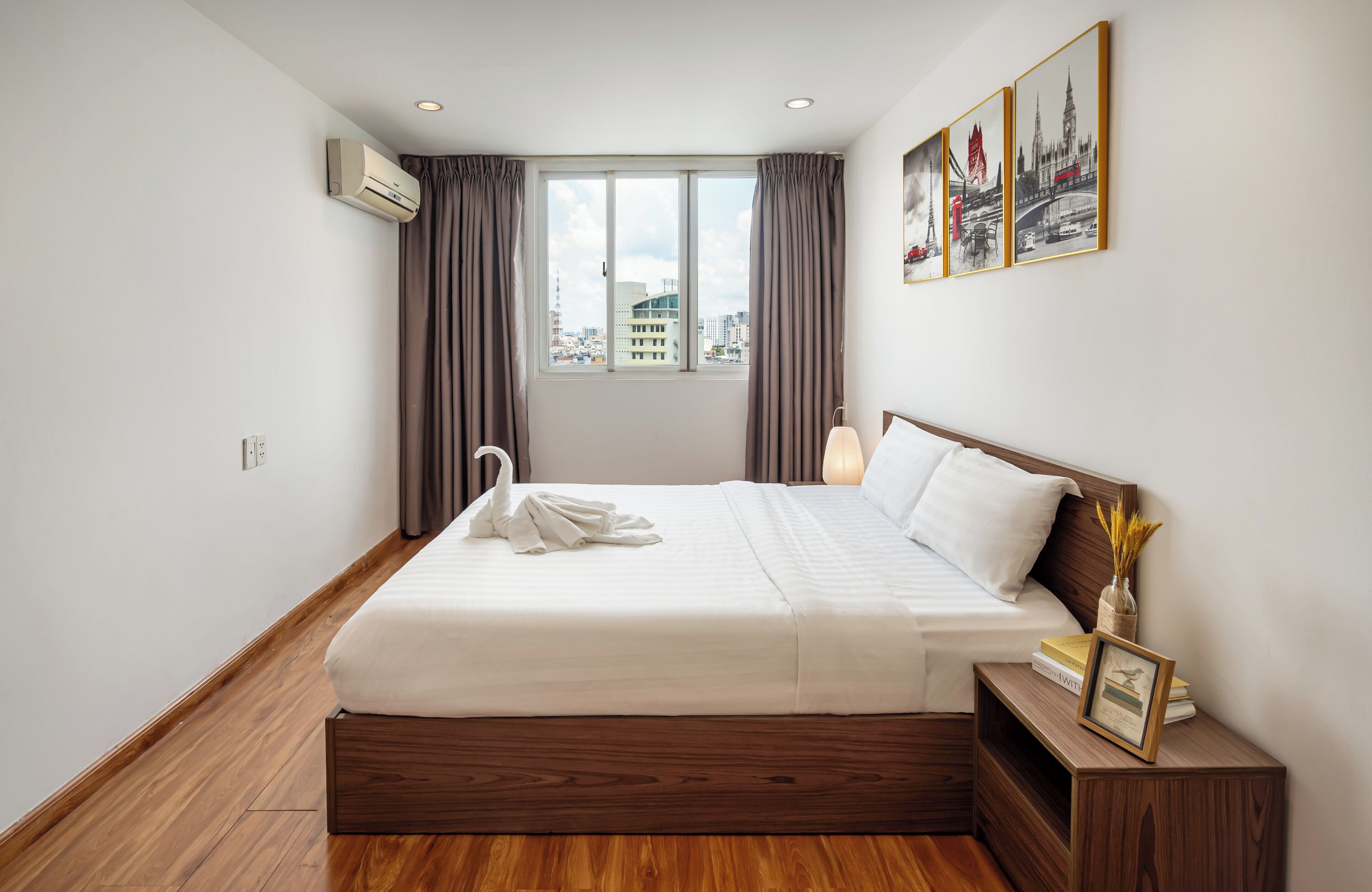 A hotel room with a large bed, a nightstand, a bedside lamp, a window with curtains, and a city view.