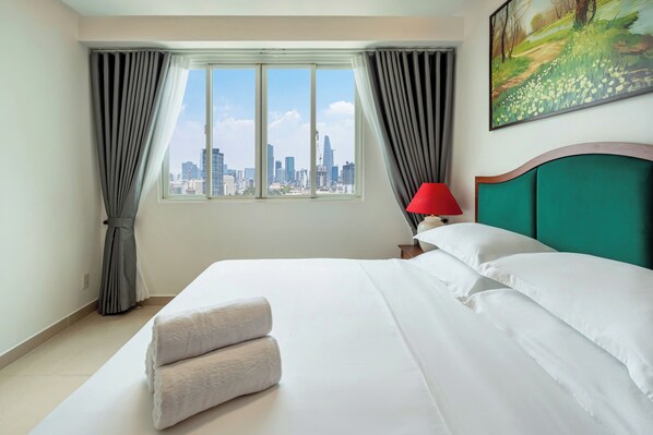 A hotel room with a large bed, a view of the city skyline, a red lamp, and a painting on the wall.