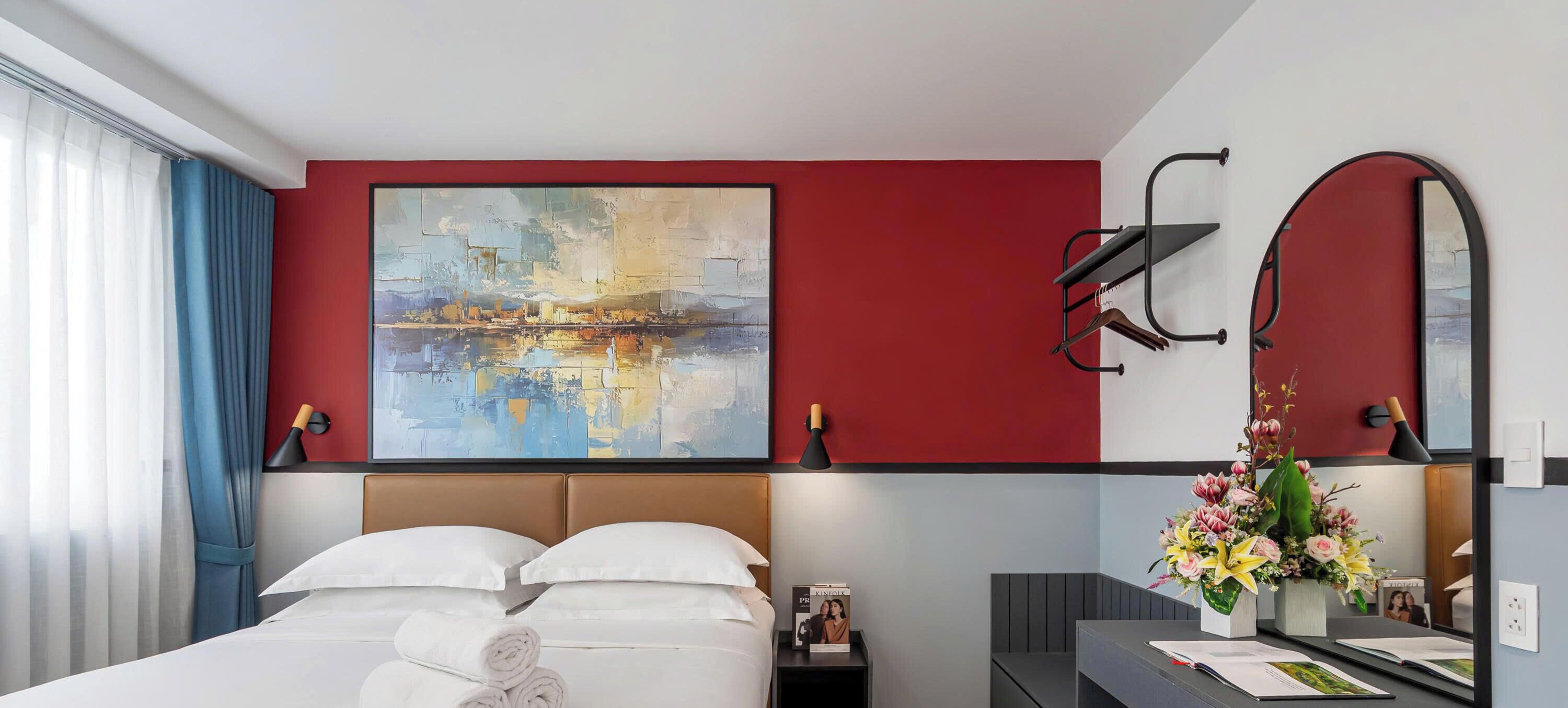 Une chambre d’hôtel avec un lit, un grand tableau abstrait, un miroir, un bureau agrémenté d’un vase de fleurs et une table de chevet avec une lampe.