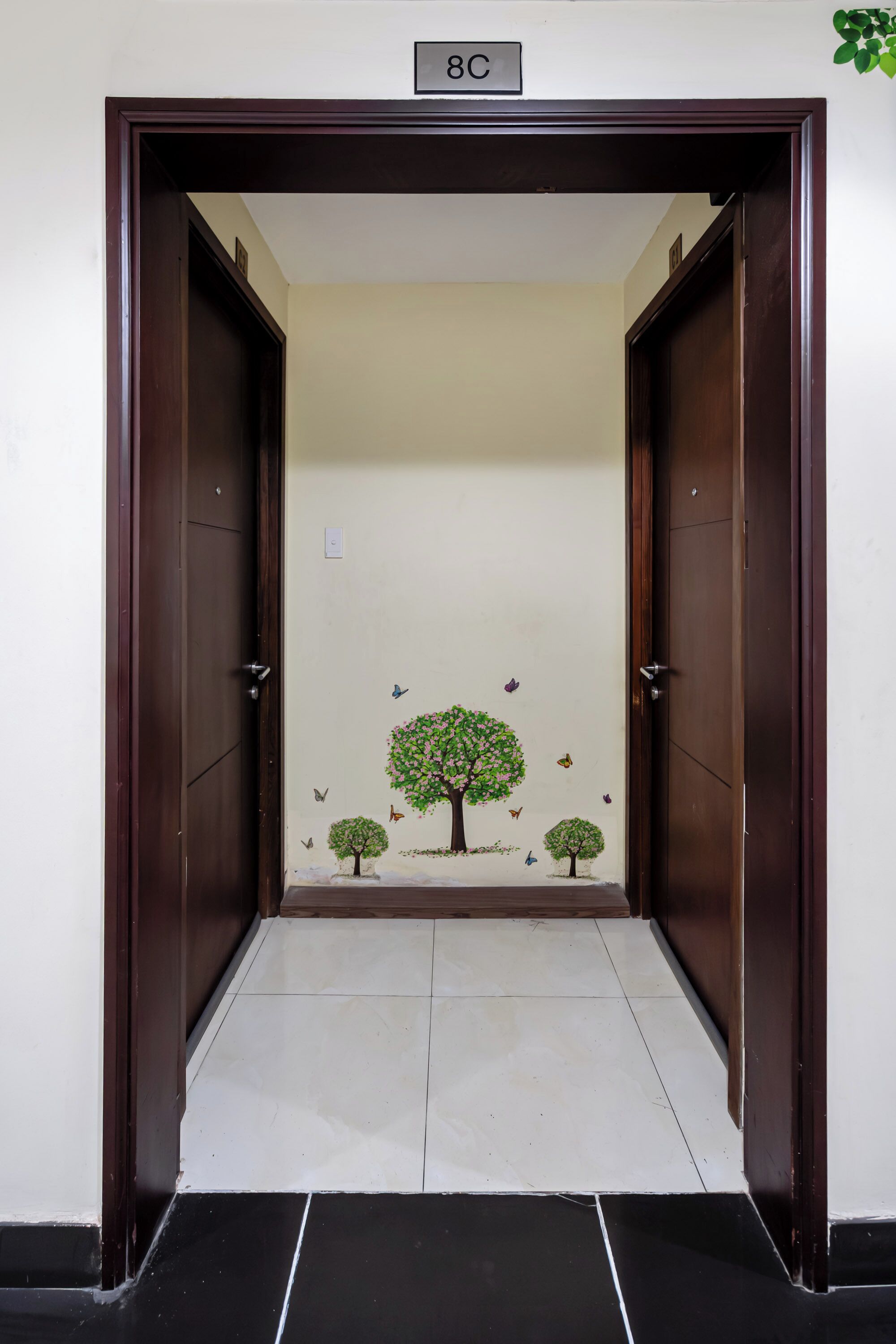A hallway with a door marked '8C,' a tiled floor, and a wall with a tree and butterfly decoration.