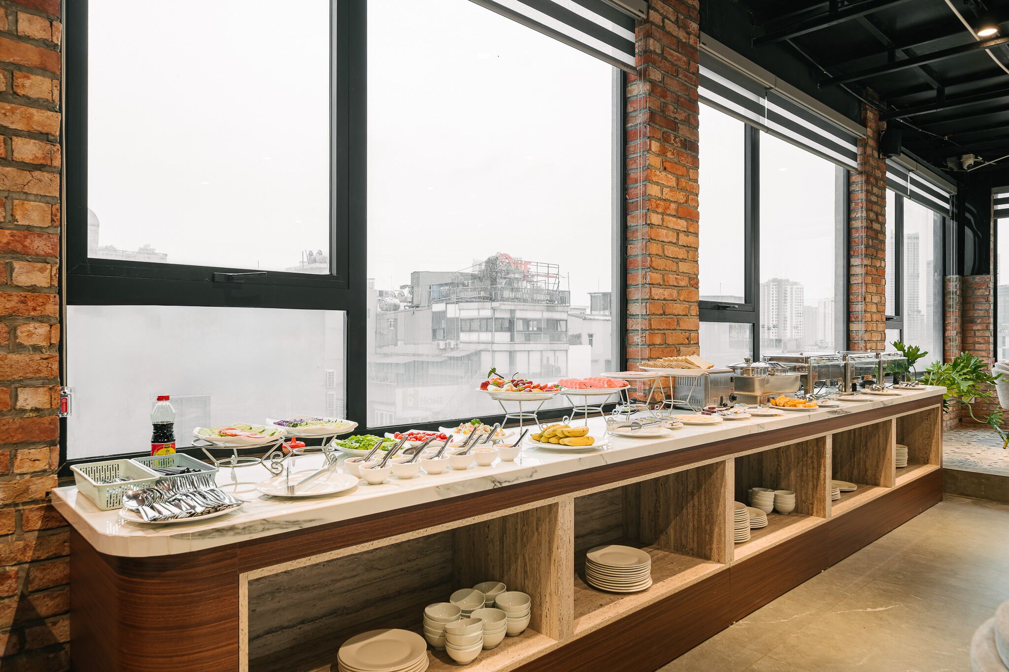 A buffet setup with various food items and utensils, large windows with a city view, and brick walls.