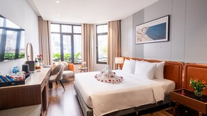 A hotel room with a large bed, a desk with a chair, a small table with a vase of flowers, and a large window with curtains. - San Seize Hanoi Hotel (Hanoi)
