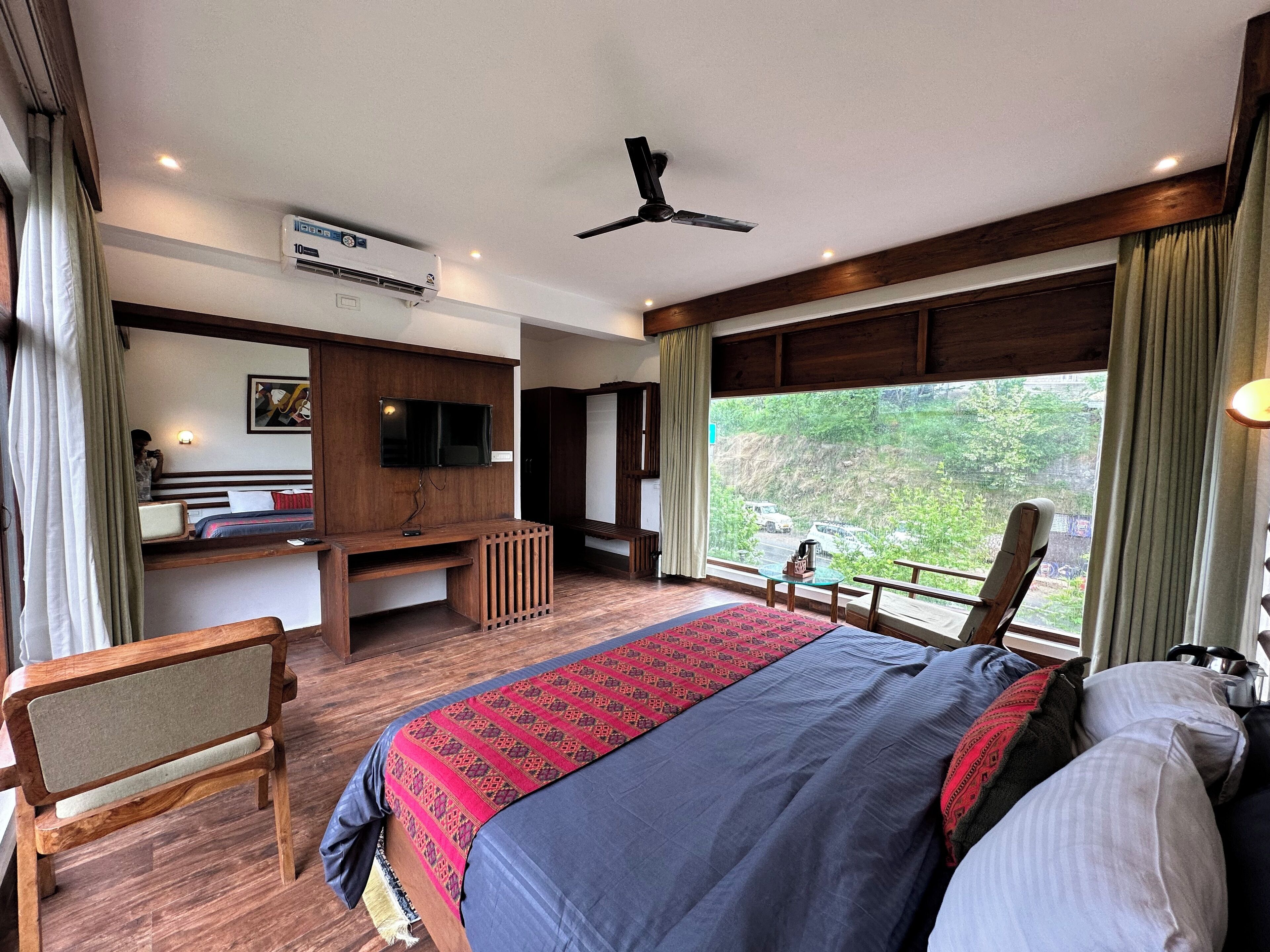 A hotel room with a large bed, a TV mounted on the wall, a desk with a chair, and a window with a view of greenery.