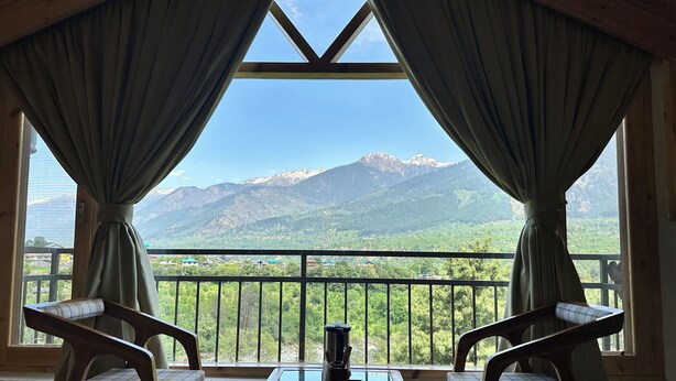 A room with a view of mountains through large windows, two chairs, and a table. - Crimson inn by Senderos De Pine (Kullu)
