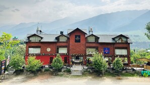 A two-story building with a red facade and multiple windows, surrounded by greenery and mountains in the background. - Crimson inn by Senderos De Pine (Kullu)