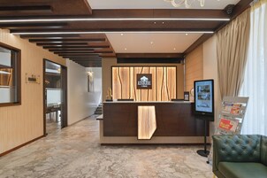 Uma área de recepção de hotel com um balcão de madeira, uma tela digital e uma área de estar.