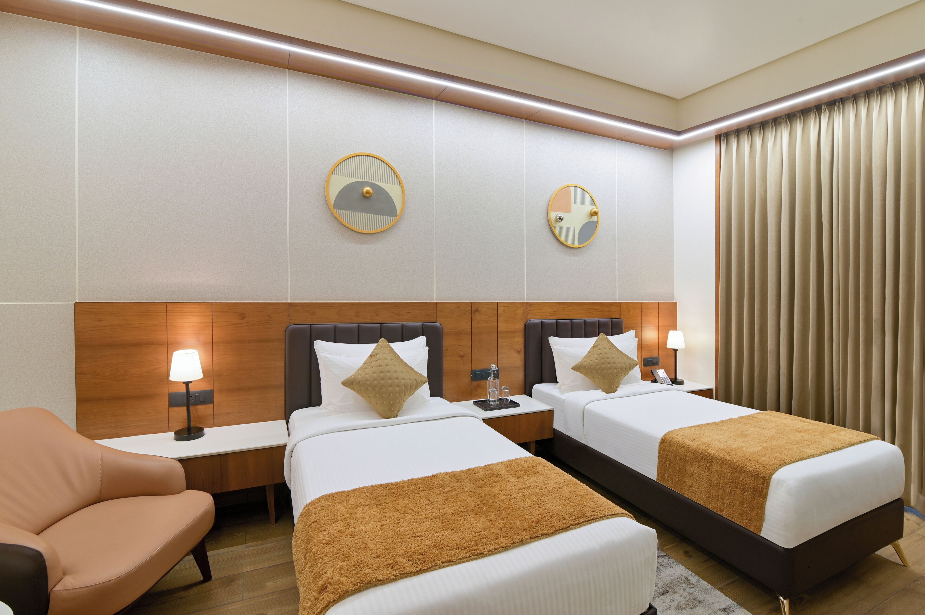 Habitación de hotel con dos camas, una mesita de noche con lámpara, una silla y un ventanal con cortinas.